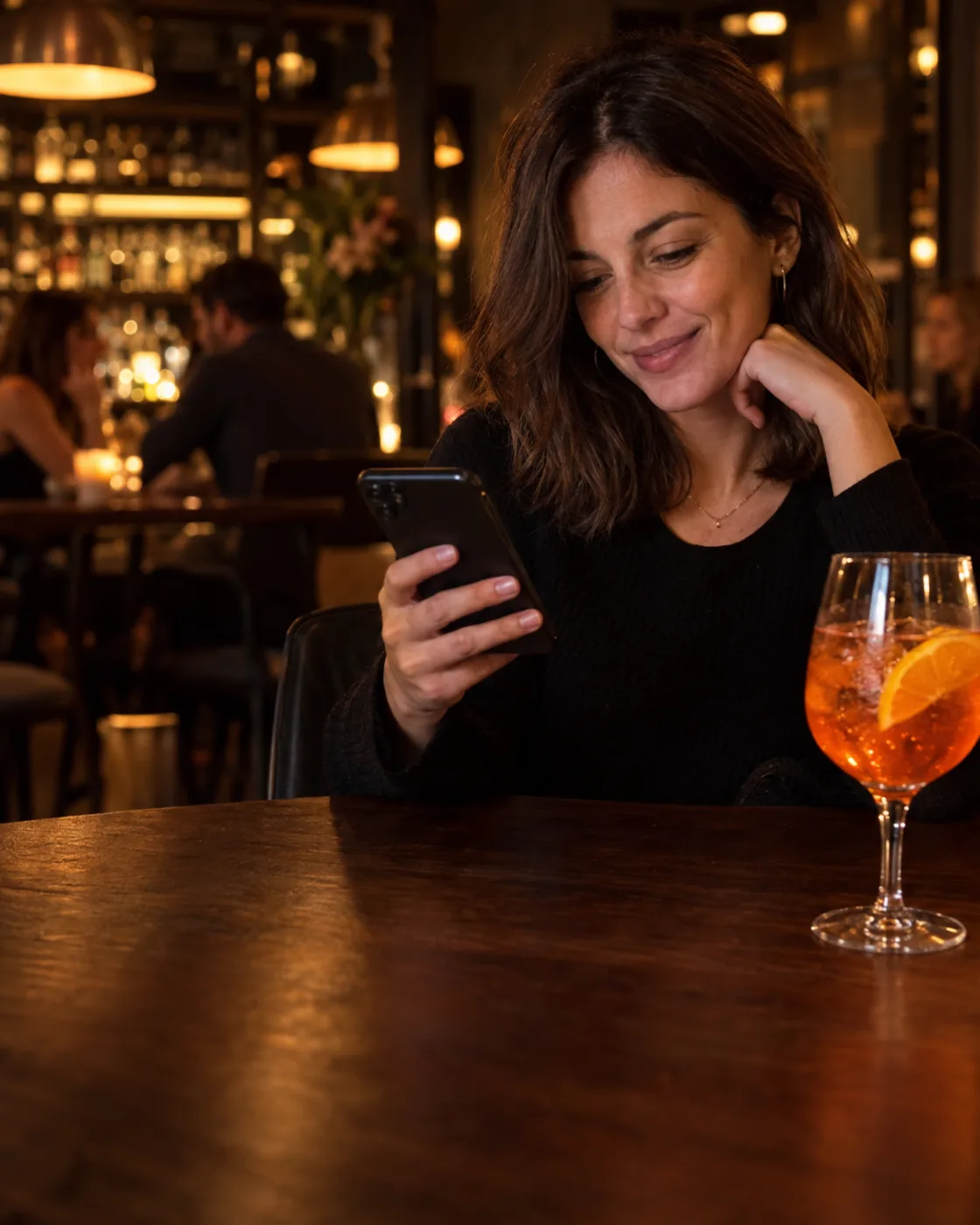 Una cliente consulta lo smartphone al tavolo di un cocktail bar, accanto a uno Spritz.